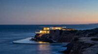 Modern glass house perched on a rocky coastline at dusk, its warm interiors glowing over the calm sea.