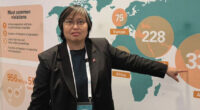 Woman in a black blazer points at a world map infographic during a presentation or conference.