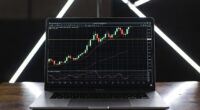 Laptop on a wooden desk displaying a candlestick stock chart with moving average lines and indicators on screen, against a dark background with illuminated bars behind.