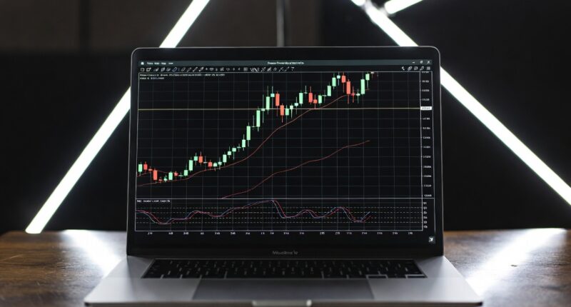 Laptop on a wooden desk displaying a candlestick stock chart with moving average lines and indicators on screen, against a dark background with illuminated bars behind.